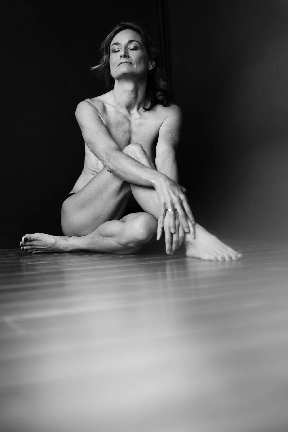A black and white photo of a nude woman sitting on the floor with her eyes closed, arms crossed over her knees, against a plain dark background.