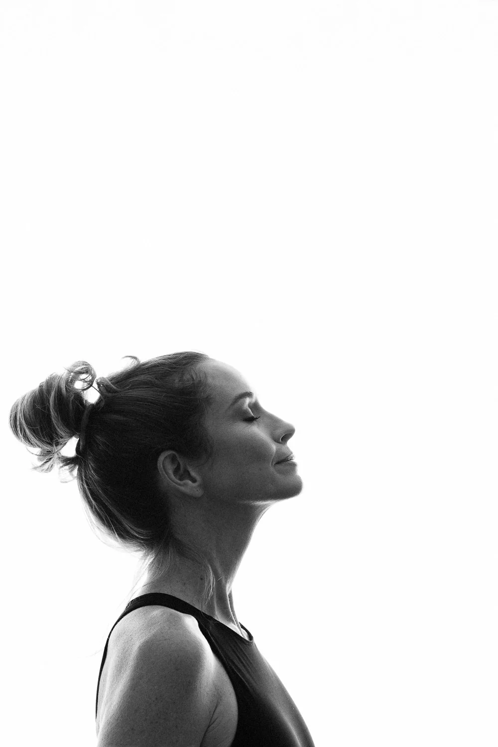 Side profile of a woman with her eyes closed, smiling faintly, wearing a tank top, with her hair tied in a bun, against a plain white background.