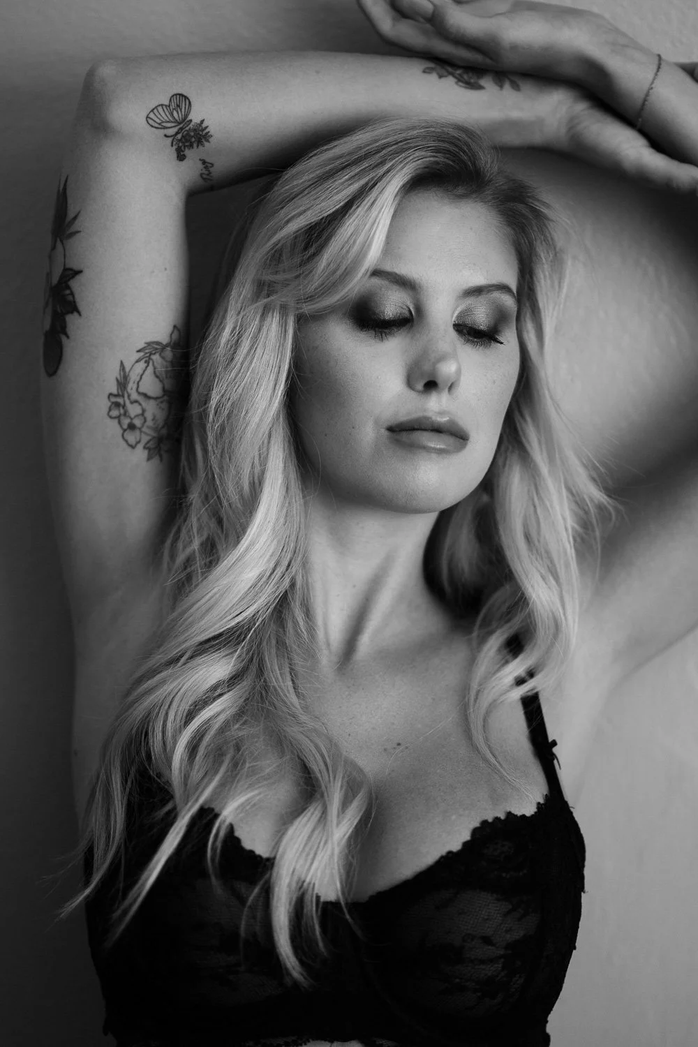 Black and white portrait of a woman with closed eyes, light wavy hair, tattoos on her arm, wearing lace lingerie, with her arms raised above her head.
