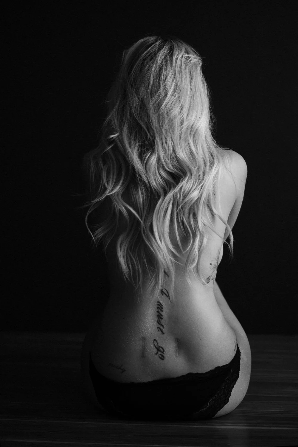 Black and white photo of a woman with long, wavy hair sitting with her back to the camera, wearing black lace underwear, with tattoos visible on her lower back and side.