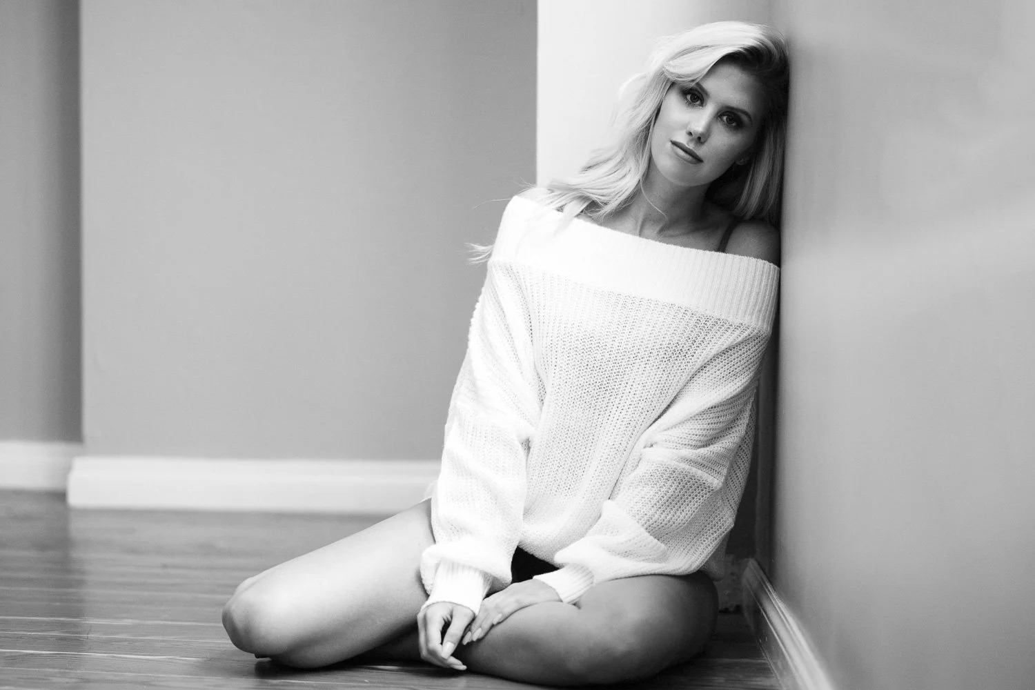 Black and white photo of a woman with long blonde hair, sitting on the floor, leaning against a wall, wearing an off-the-shoulder sweater.