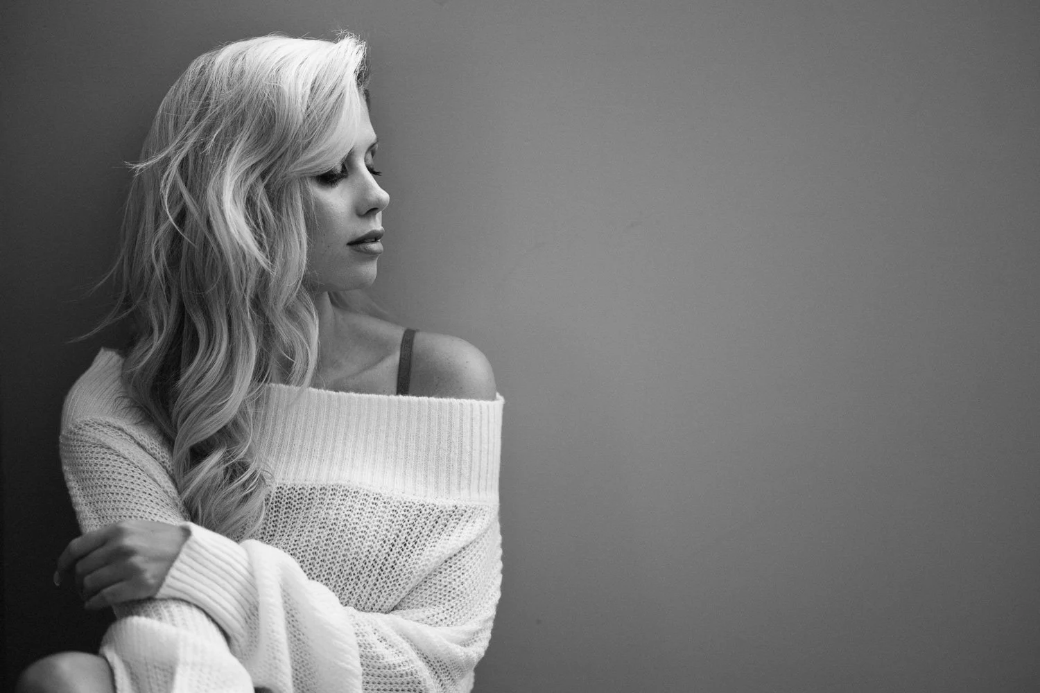 A black and white photo of a woman with long, wavy hair, wearing a off-the-shoulder sweater, looking to the left against a plain wall.