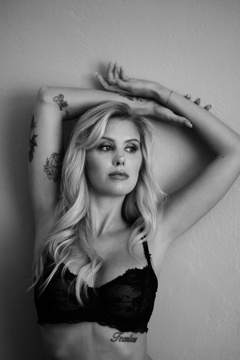 A black and white photo of a woman with long wavy blonde hair, tattoos on her arms, wearing a black lace bra, with her arms raised above her head and looking to the side.