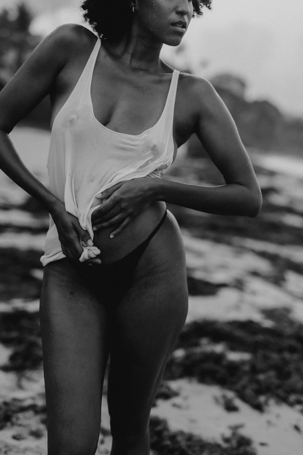 Black and white photo of a woman wearing a wet tank top and underwear, standing outdoors with a blurred natural background.