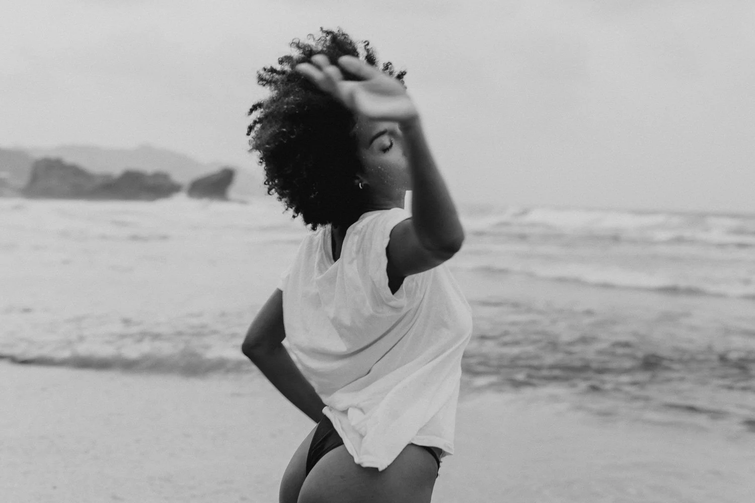 Black and white photo of a woman with curly hair on a beach, wearing a loose shirt and underwear, with an arm raised and hand on her head, ocean waves in the background