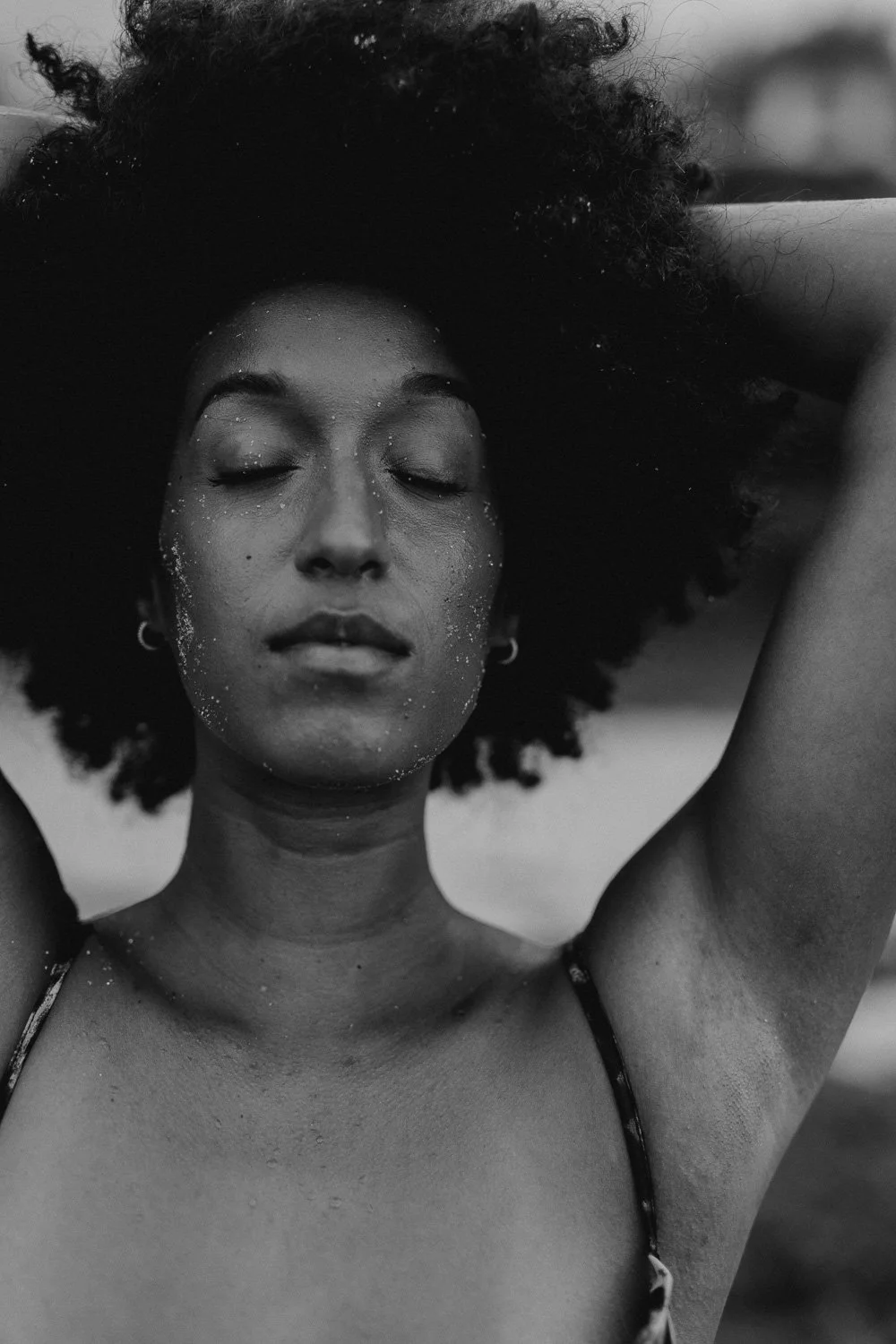 A woman with natural curly hair and closed eyes, with water droplets on her face and shoulders, posing with her arms raised behind her head in a black and white photograph.