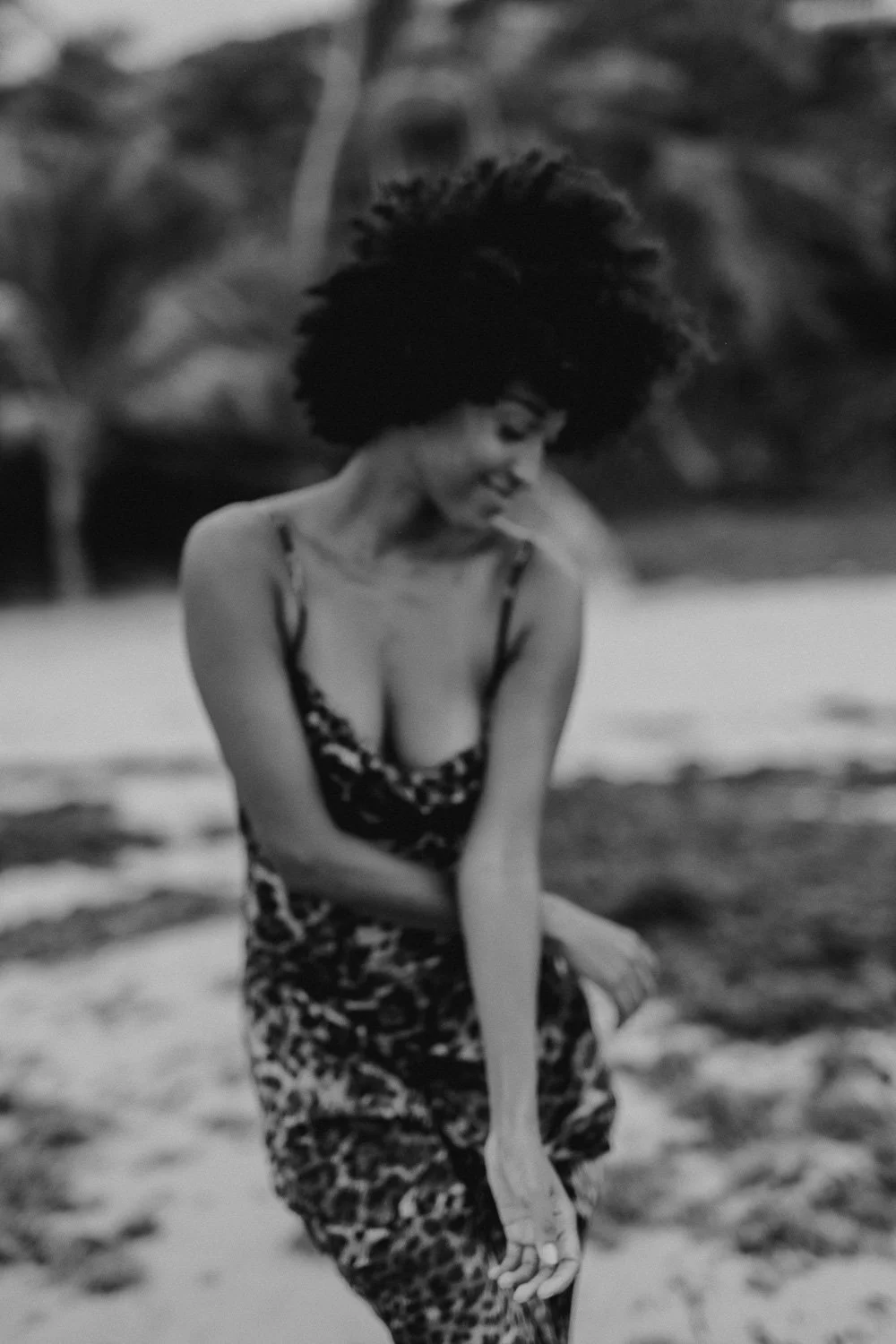 A woman with curly hair wearing a patterned dress, standing outdoors with trees in the background, smiling and looking down.