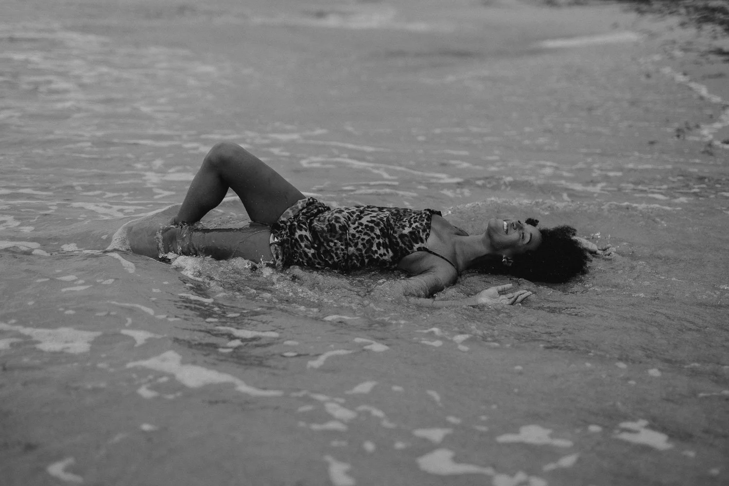 A woman lying on her back on the beach, smiling, with her head resting on the sand and waves washing over her. She is wearing leopard print shorts and a spaghetti strap top.