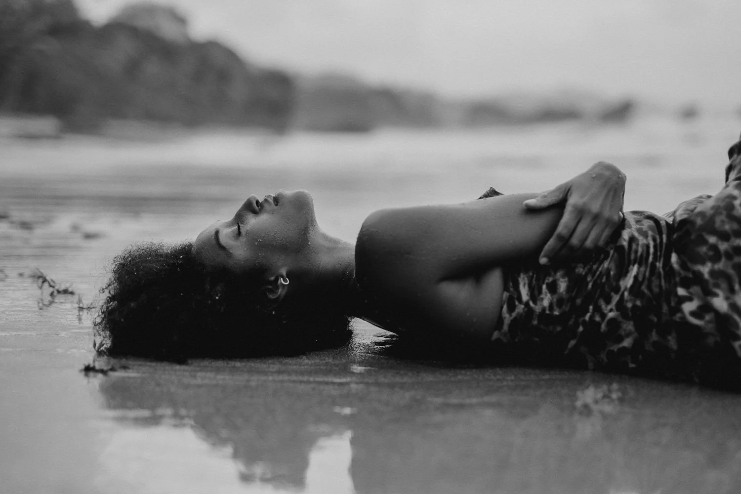 A woman lying on a beach with her eyes closed, arms crossed over her chest, and her head resting on the sand. She has curly hair and appears relaxed. The background shows a blurred shoreline and distant rocks.