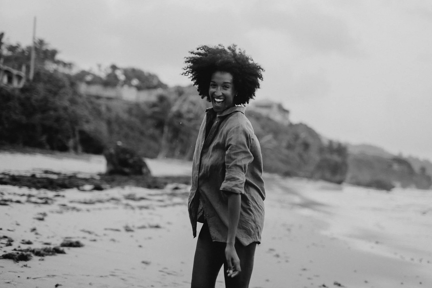 A smiling woman with curly hair standing on the beach, wearing a long shirt or jacket, with cliffs and trees in the background.