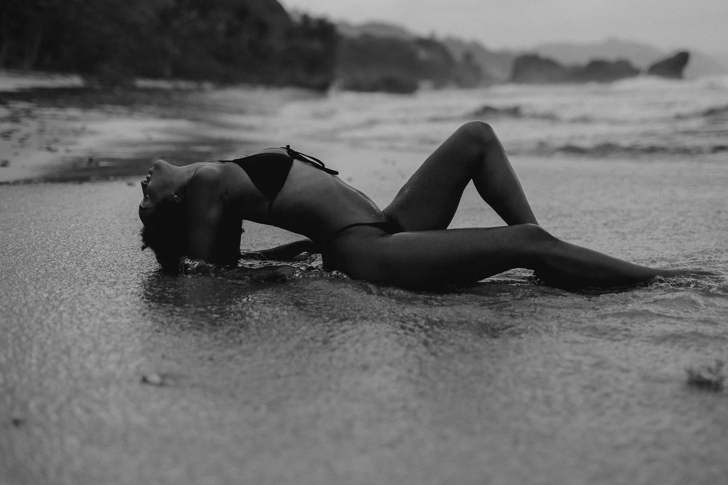 A woman in a bikini lying on a beach with her head tilted back and her eyes closed, with waves and cliffs in the background.