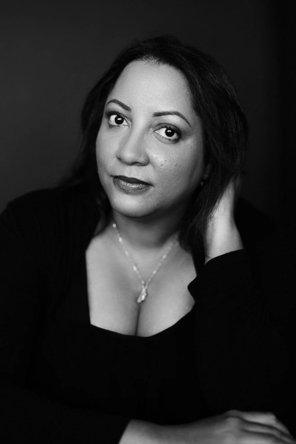 Black and white portrait of a woman with dark hair, wearing a necklace, looking at the camera, with one hand resting on her neck.