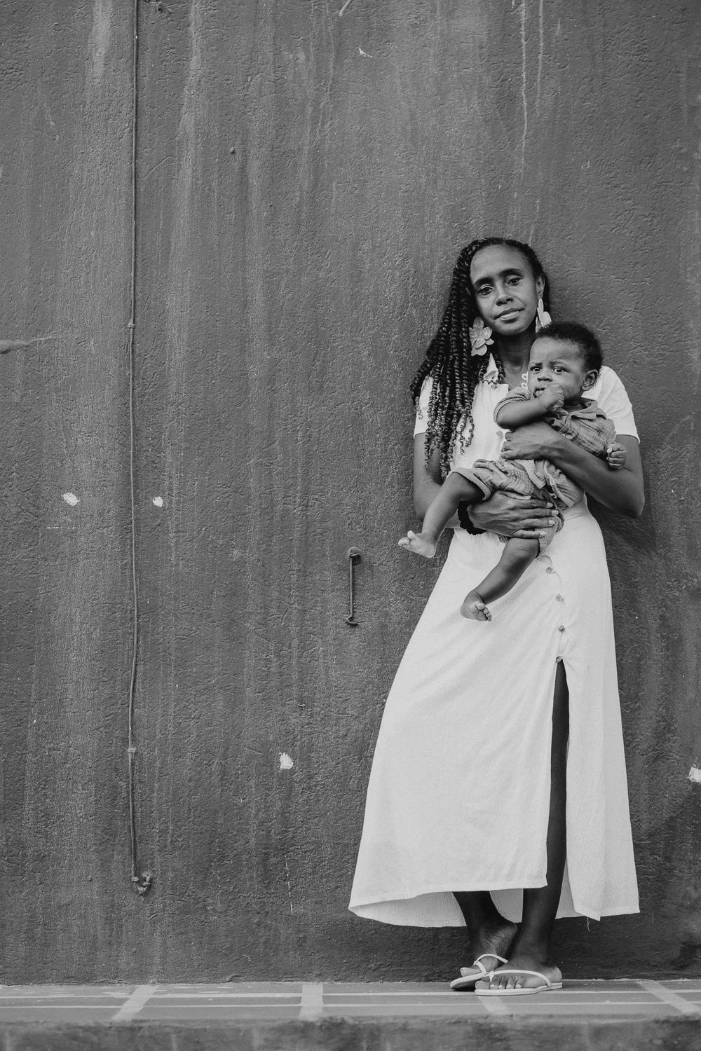A woman with long braided hair holding a young child, standing against a textured wall.