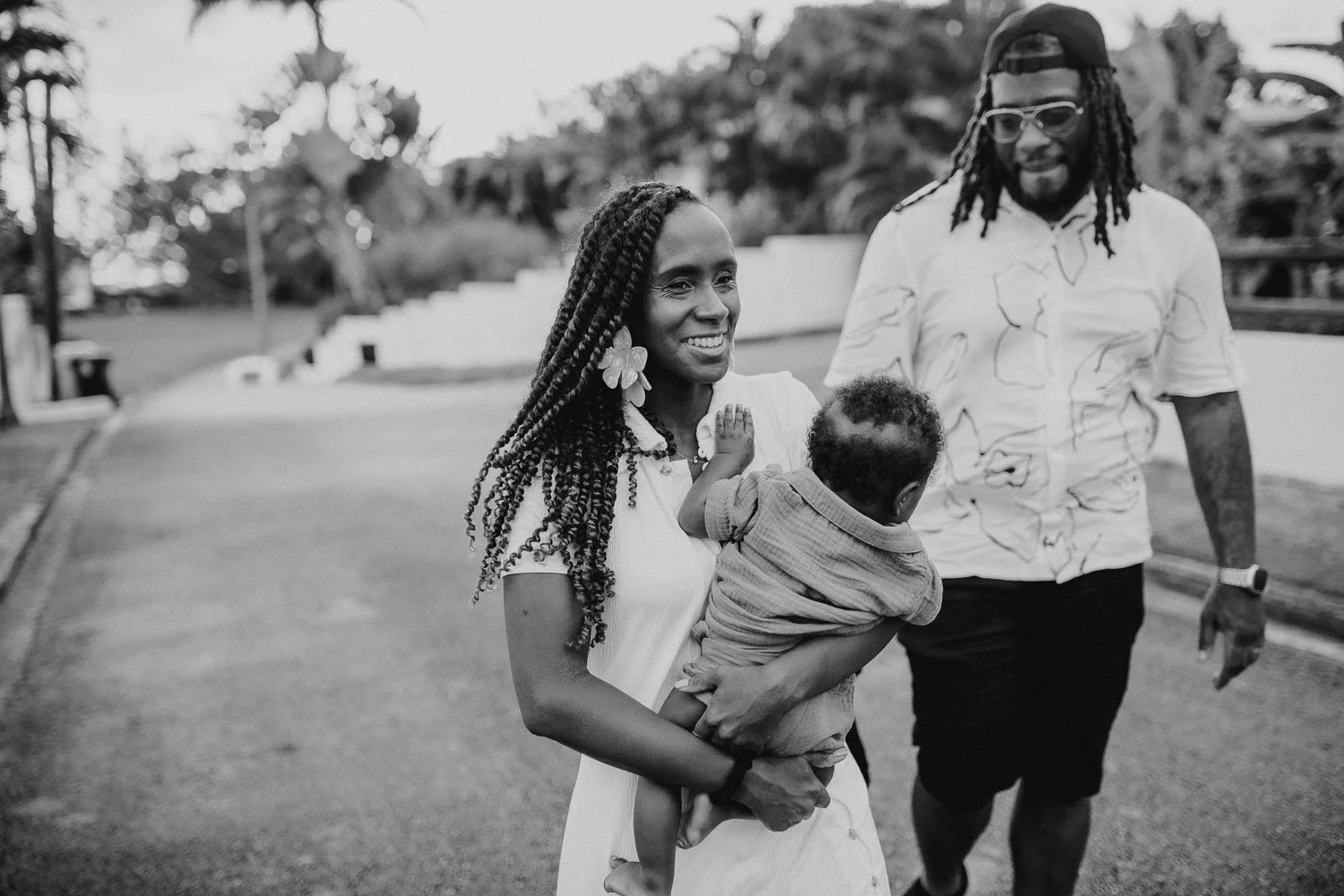 A woman with braided hair and a flower earring holds a young child, smiling, while a man with dreadlocks, sunglasses, and a patterned shirt walks beside them in a park-like setting.