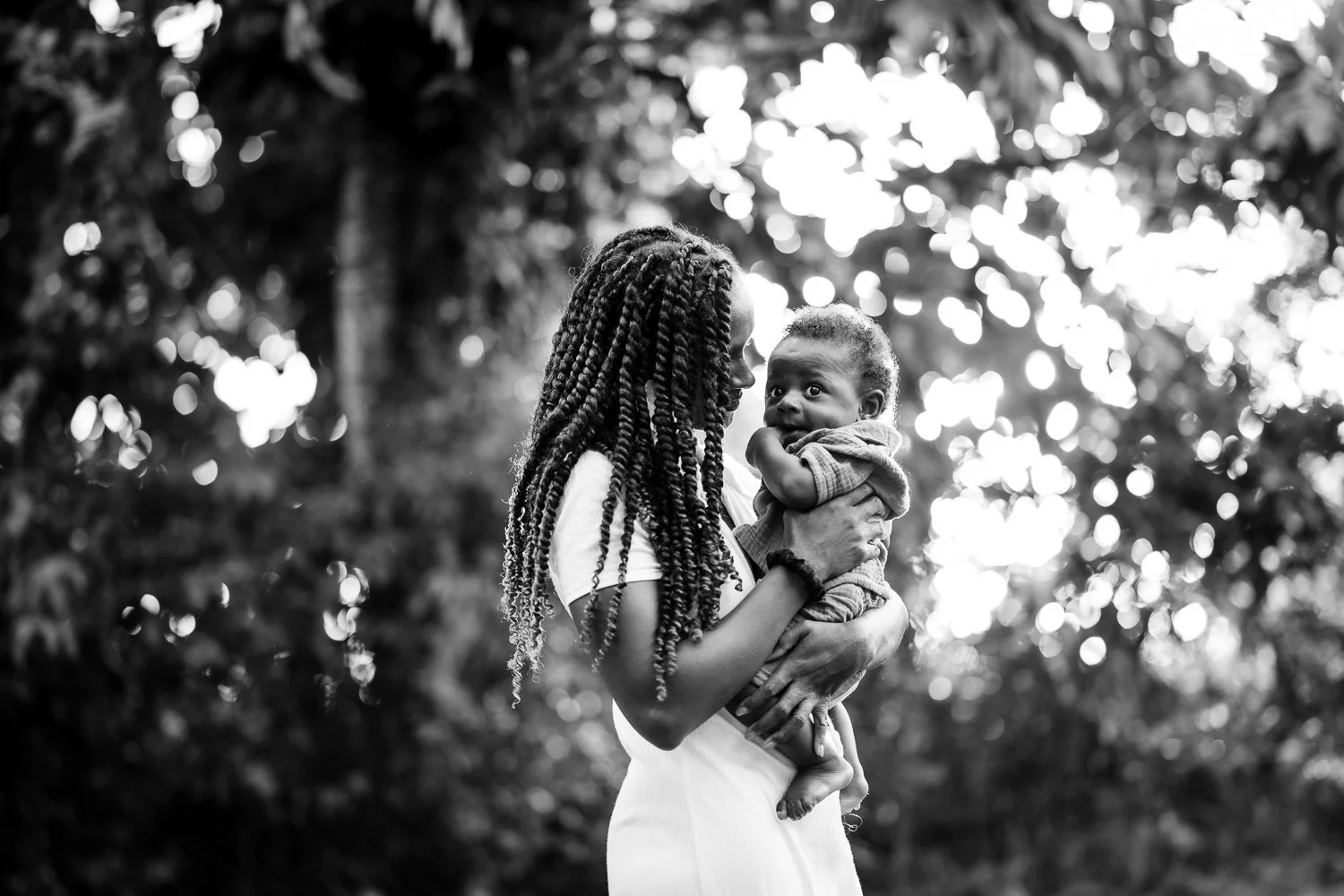 A woman with long braided hair holding a baby outdoors, with a background of trees and blurred sunlight.