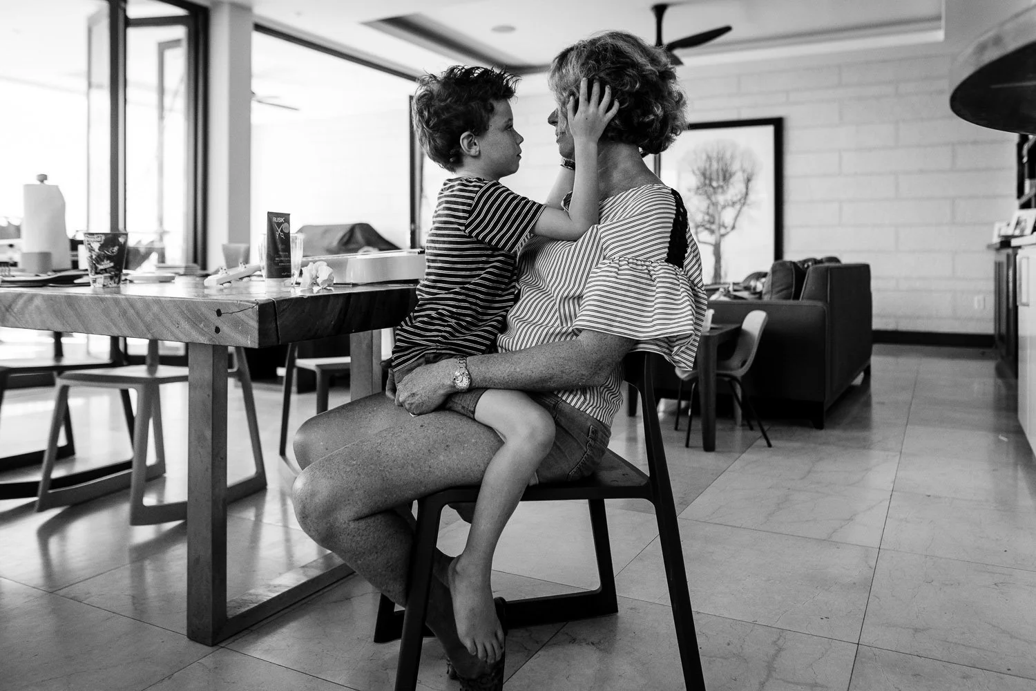 A young boy and an elderly woman sharing a tender moment, with the boy sitting on the woman's lap, touching her face, in a modern, well-lit dining area.