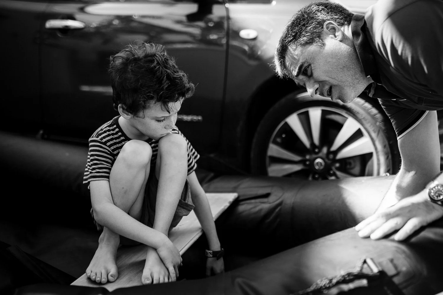 A young boy is sitting on the edge of a boat, bent forward, with his chin resting on his knees, looking down. An adult man is leaning over the boat, looking down and speaking to the boy. Both are engaged in a serious moment. A car and a tire are visi