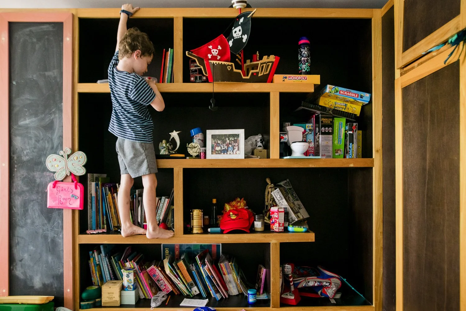 A young boy standing on a wooden bookshelf, reaching up to the top shelf. The bookshelf is filled with books, toys, and decorative items. A pink sign with a butterfly and the words 'Girl's Room' hangs on the side of the bookshelf. The background incl
