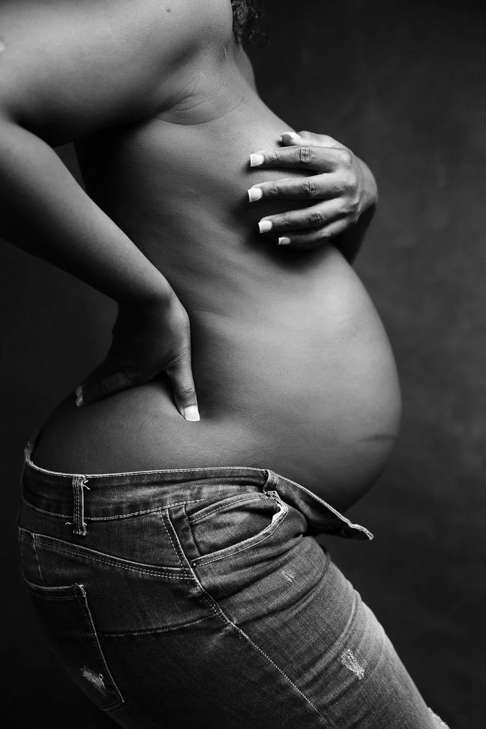 Black and white photo of a pregnant woman with one hand on her belly and the other on her side, wearing jeans.