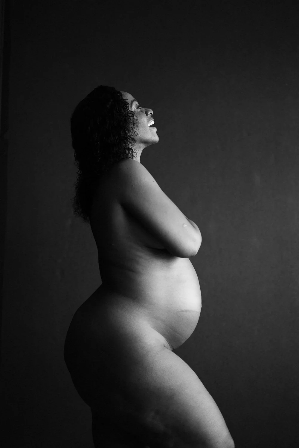 Black and white photo of a pregnant woman standing sideways with her arms crossed, smiling and looking upward, against a dark background.