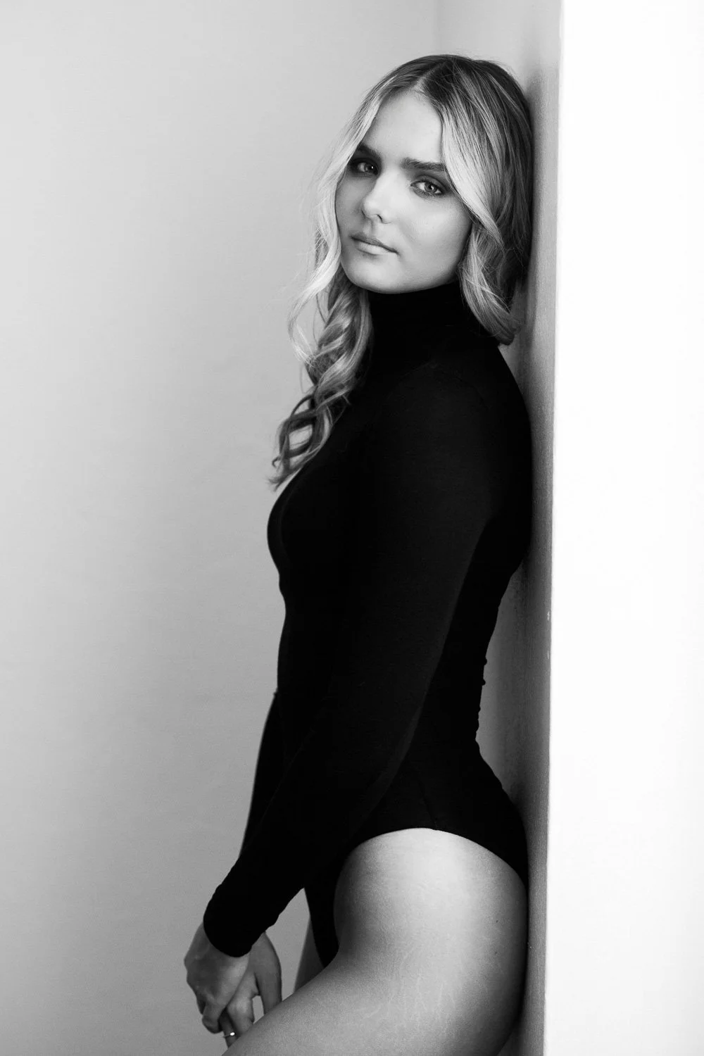 Black and white portrait of a young woman with long, wavy hair, wearing a black long-sleeve top, leaning against a corner wall.