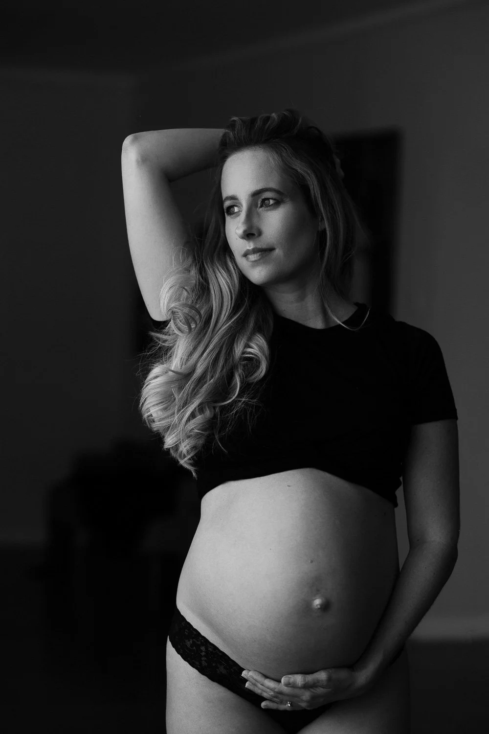 Black and white portrait of a pregnant woman with long wavy hair, wearing a black crop top and black lace underwear, holding her belly with one hand and resting her other arm on her head, in a softly lit room.