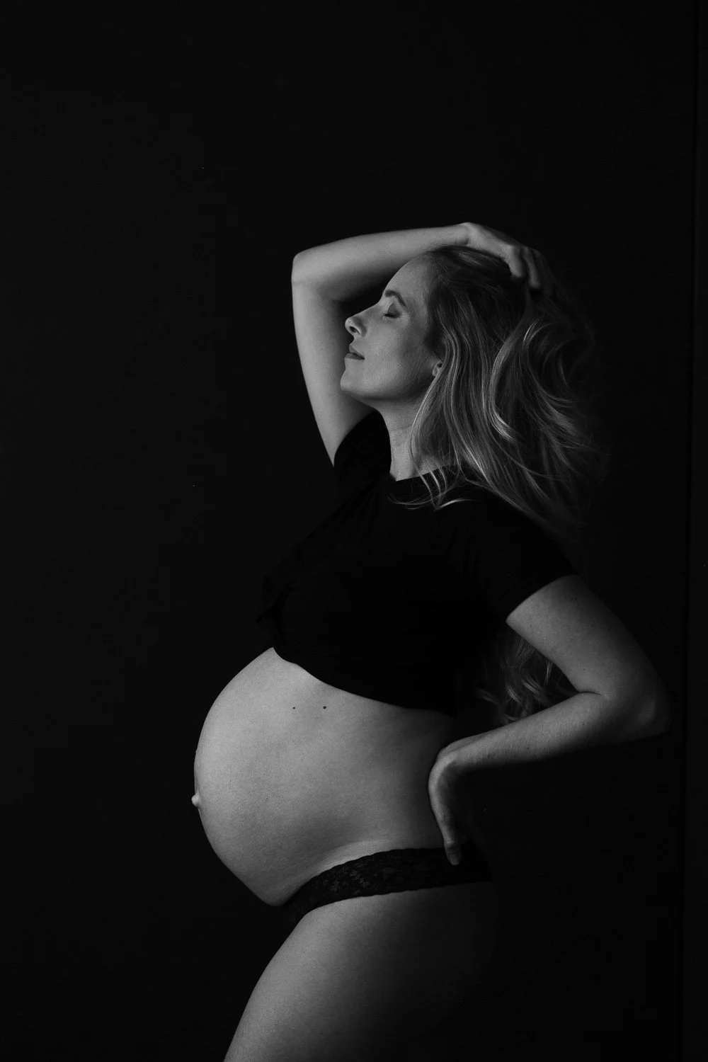 A black and white photo of a pregnant woman with long hair, her eyes closed, with her right arm raised and hand resting on her head, standing against a dark backdrop.