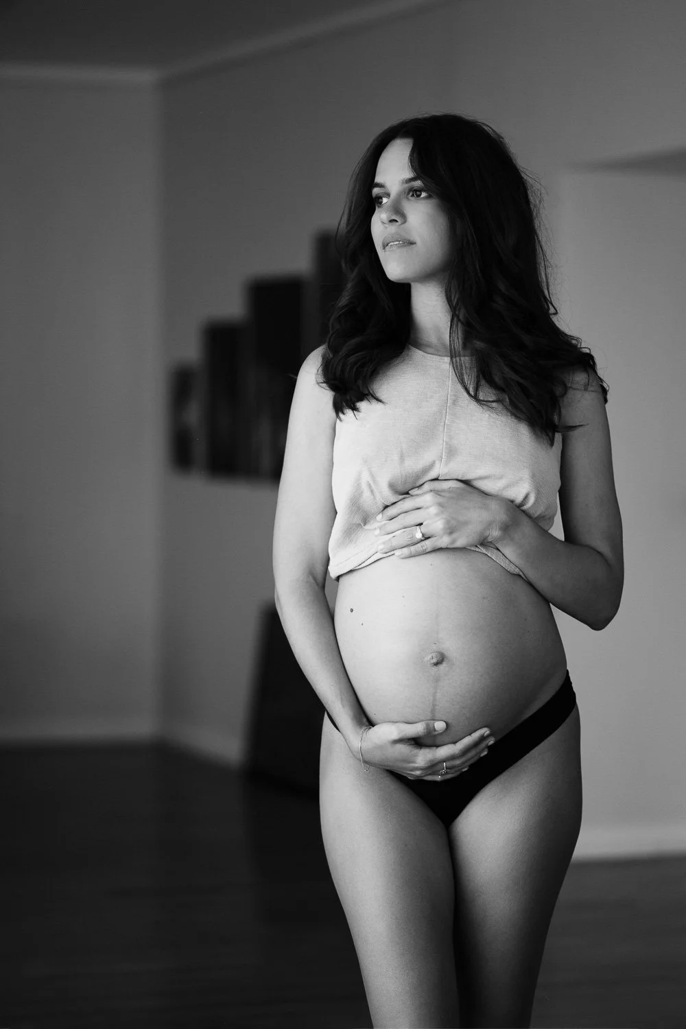 A pregnant woman with long dark hair standing indoors, wearing a sleeveless top and black underwear, gently holding her belly, looking thoughtfully into the distance.