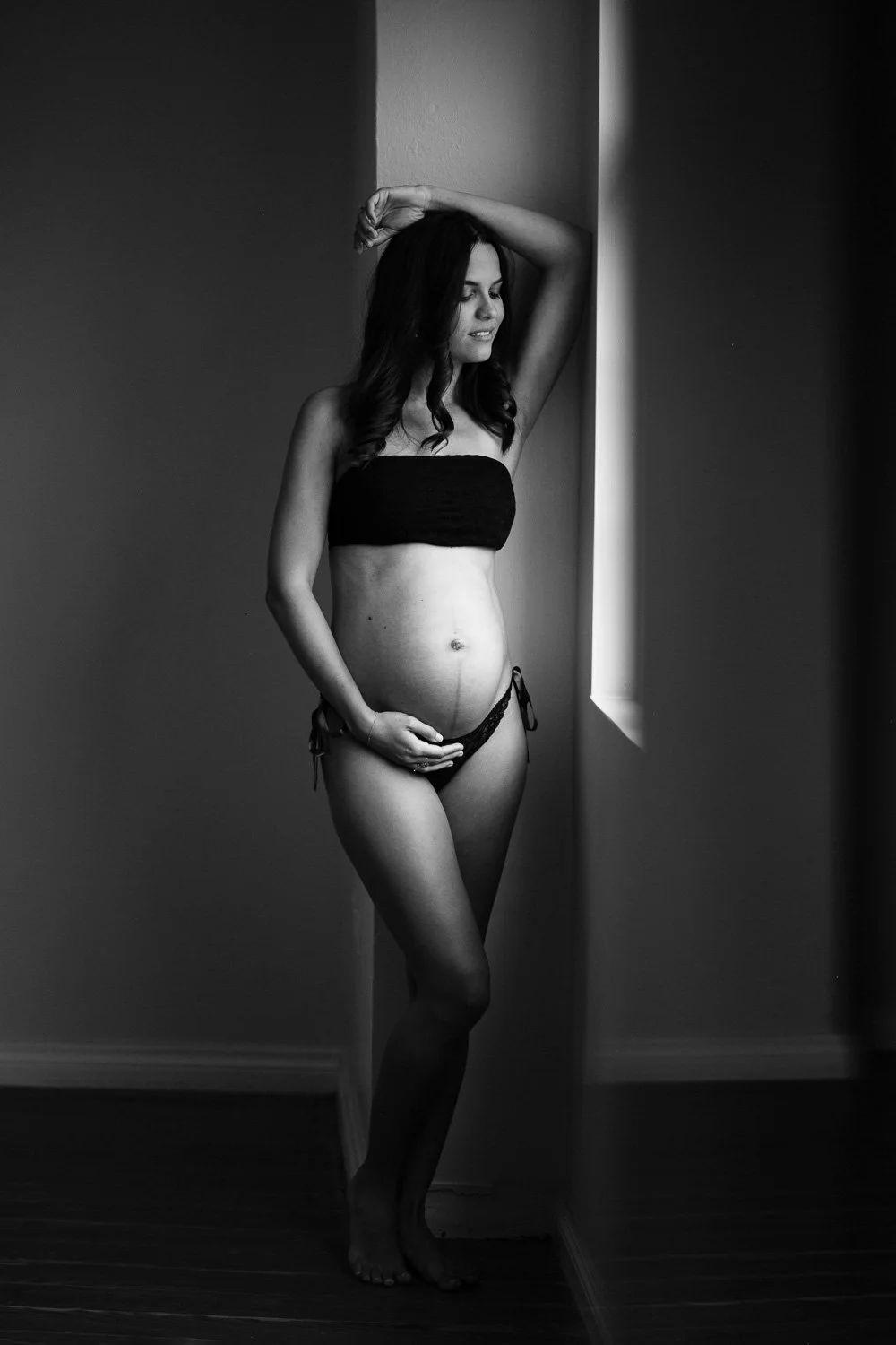 A pregnant woman standing near a window, with her left arm resting on her head and her right hand gently touching her belly, in a black-and-white photograph.