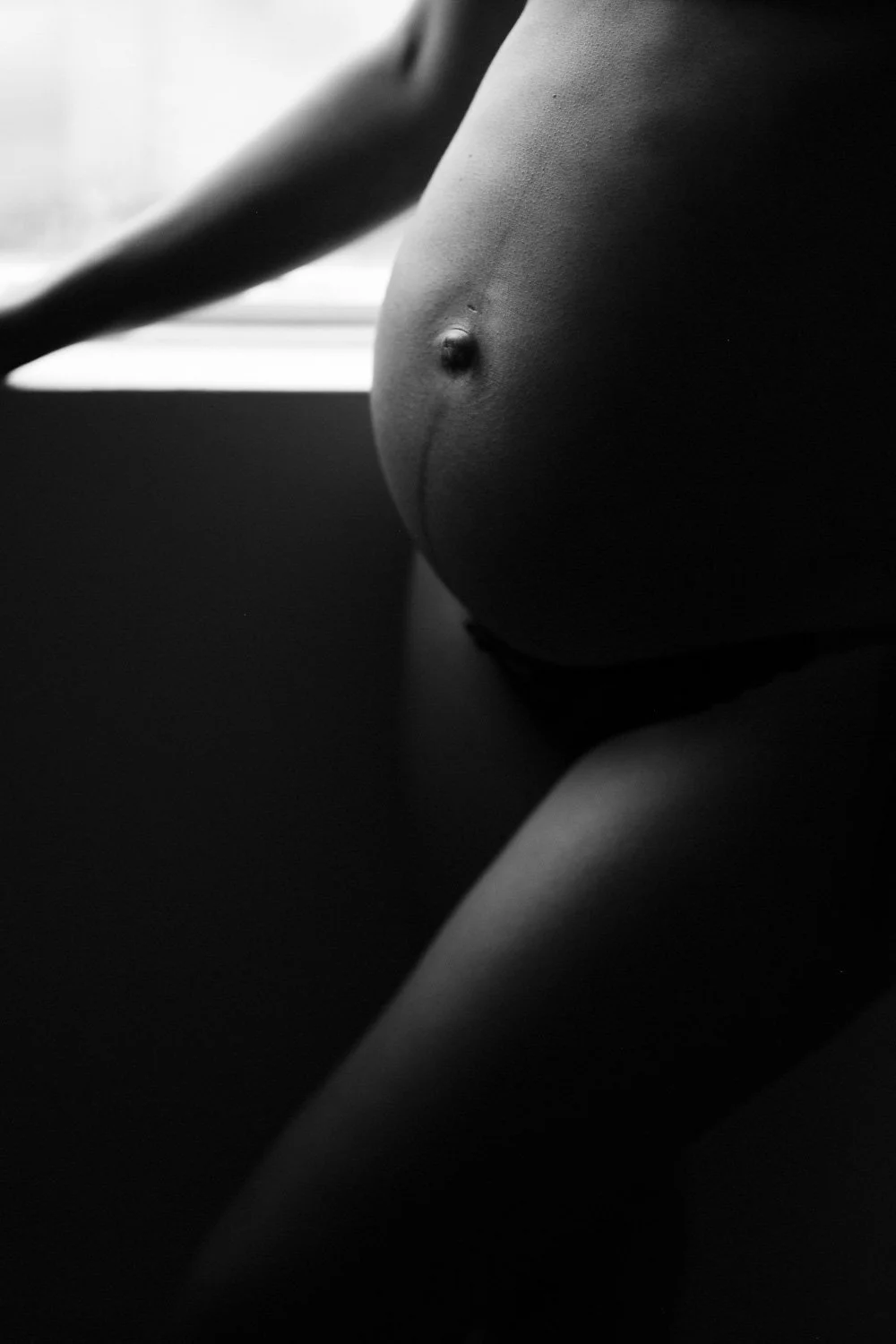 Black and white photo of a pregnant woman's torso, head not visible, with natural light highlighting her belly and arm, sitting near a window.