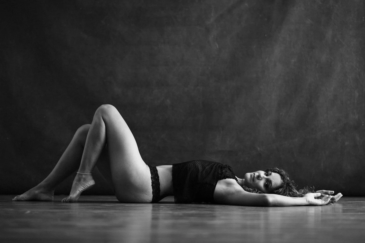 A woman with curly hair lying on a wooden floor, wearing black lace lingerie, looking directly at the camera in a black and white photo.