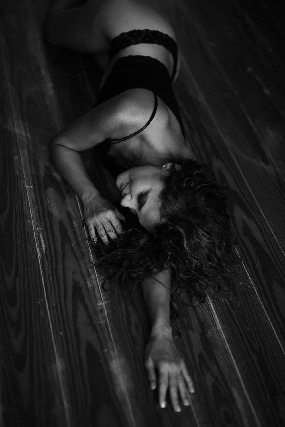A woman with curly hair lying on a wooden floor, wearing black lingerie, in a sensual pose with her eyes closed and one arm extended outward.
