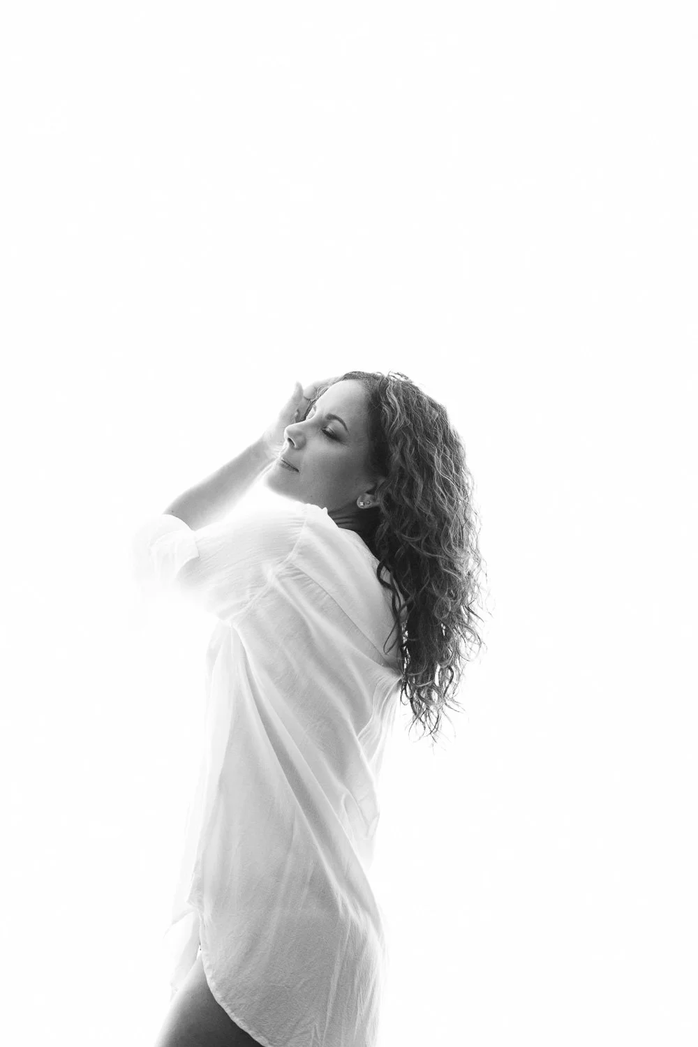 A woman with curly hair in a white shirt, posed with her eyes closed and hand on her forehead, against a bright, high-key background.