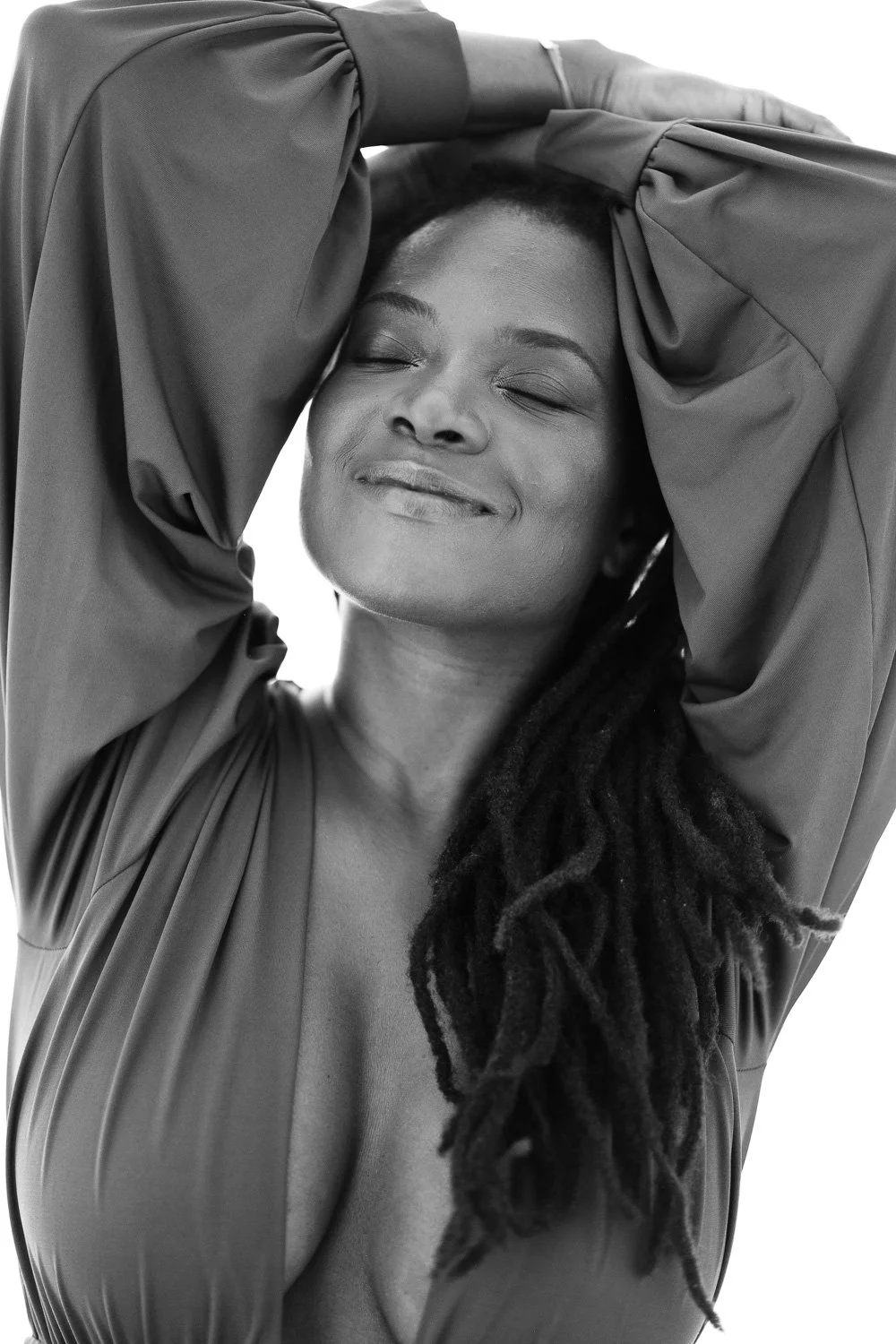 A woman with dreadlocks and closed eyes smiling with her hands resting on her head, wearing a loose long-sleeve top.