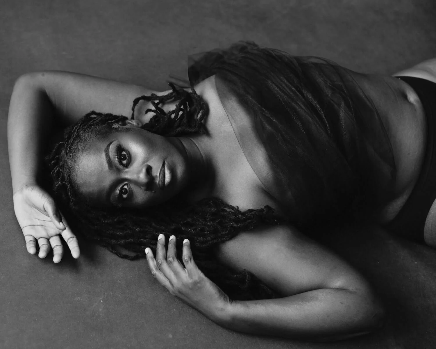 A woman with dreadlocks lying down on the floor, wearing a sheer top, looking at the camera.