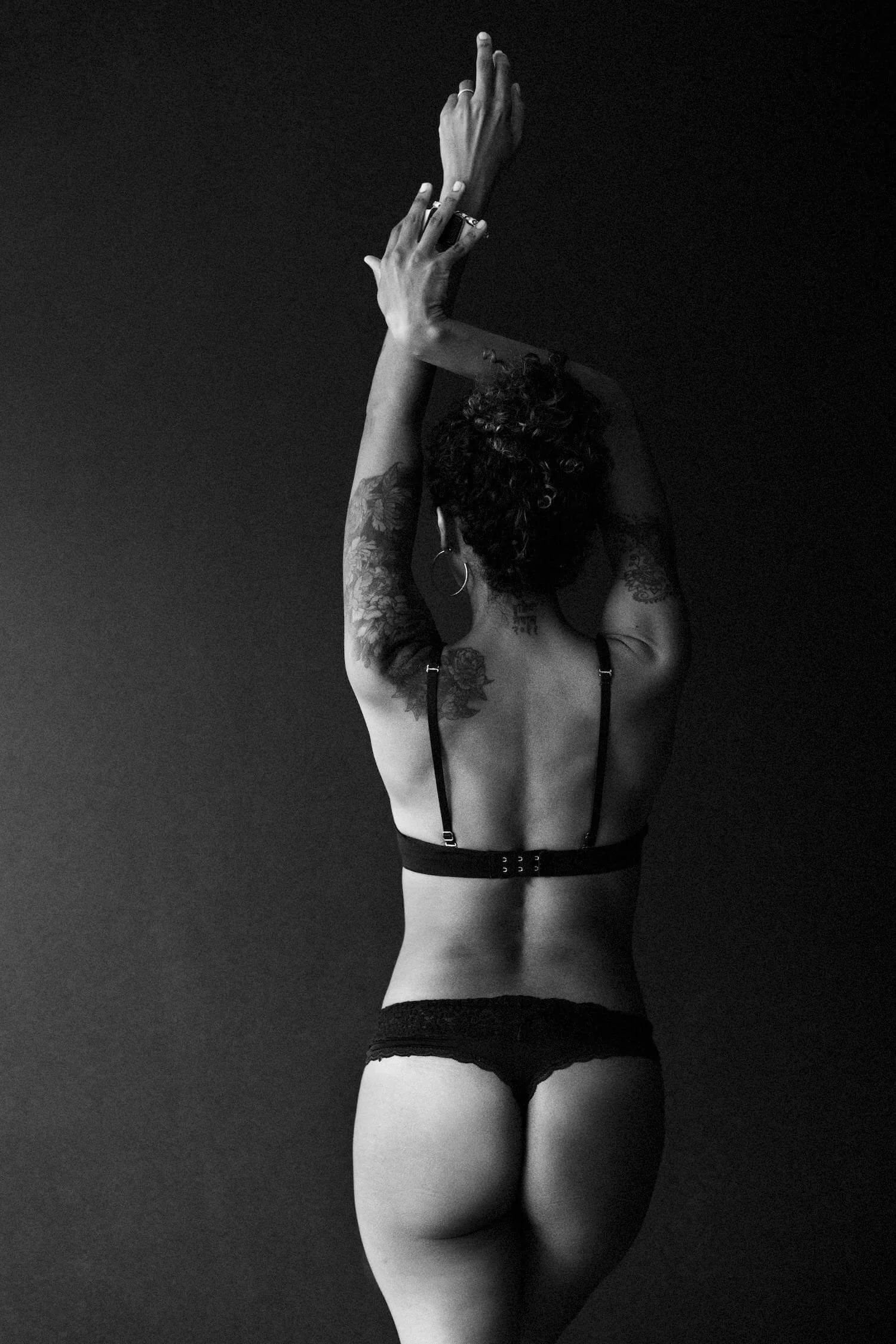 Back view of a woman with tattoos, wearing black lingerie, raising her arms with fingers pointed upwards against a plain dark background.