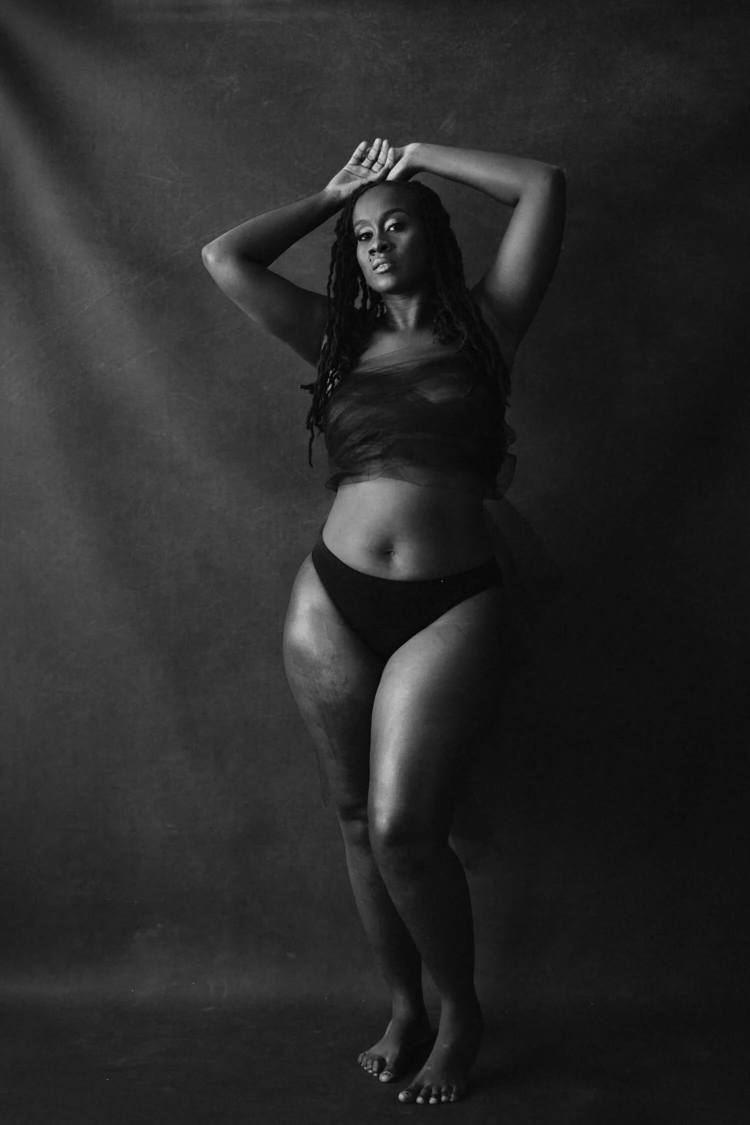 Black and white portrait of a woman with dreadlocks posing with hands above head, wearing a strapless top and underwear, standing against a textured background.