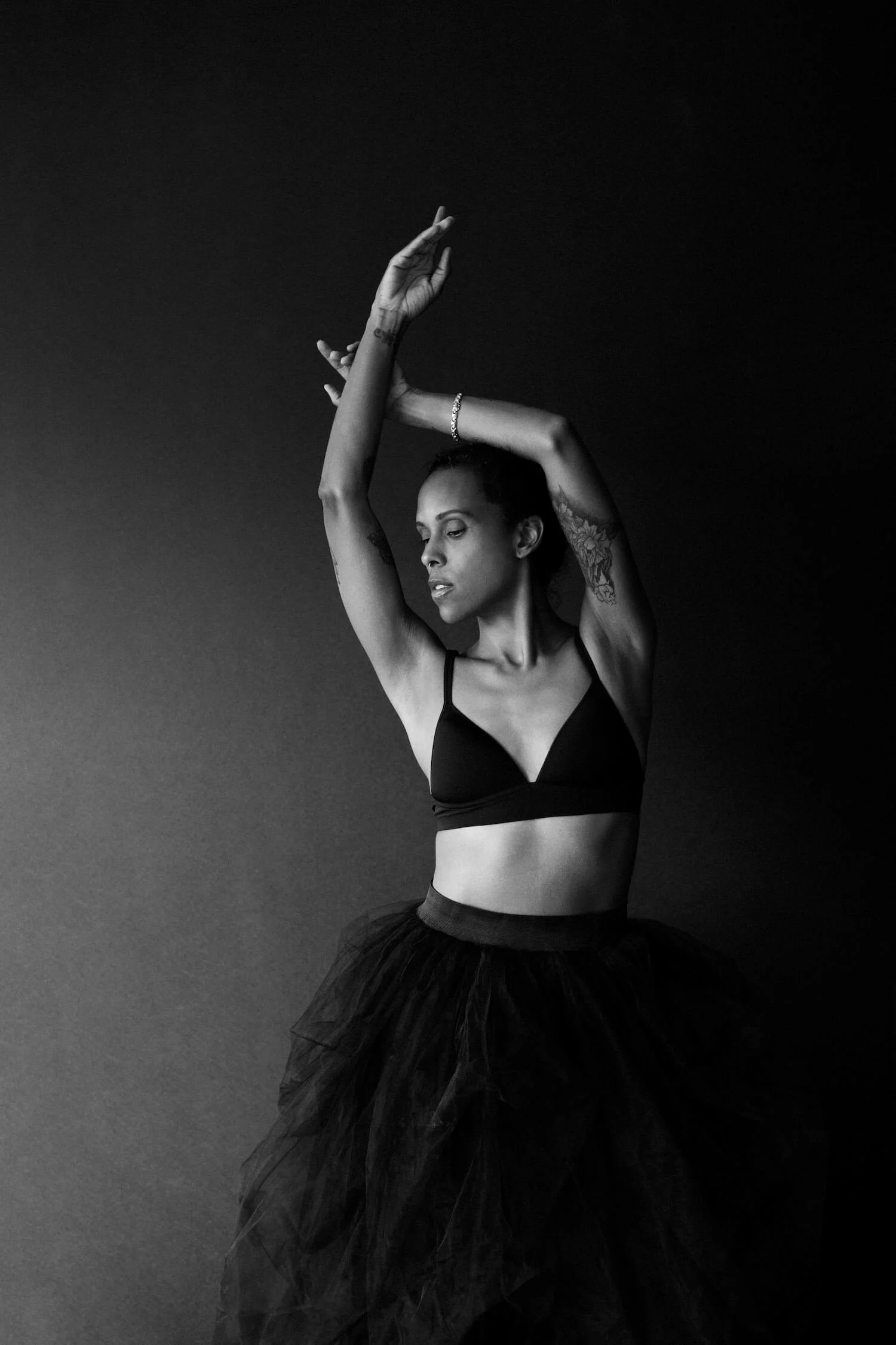 A black and white photo of a woman ballet dancer in a sports bra and tutu, with her arms raised above her head, standing against a plain dark background.