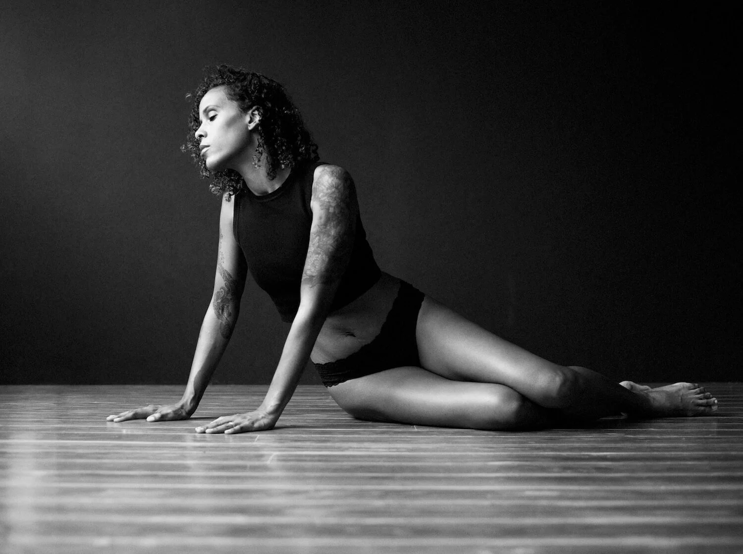A woman with curly hair in a black tank top and black shorts, kneeling on a wooden floor, facing to the left, against a dark background.