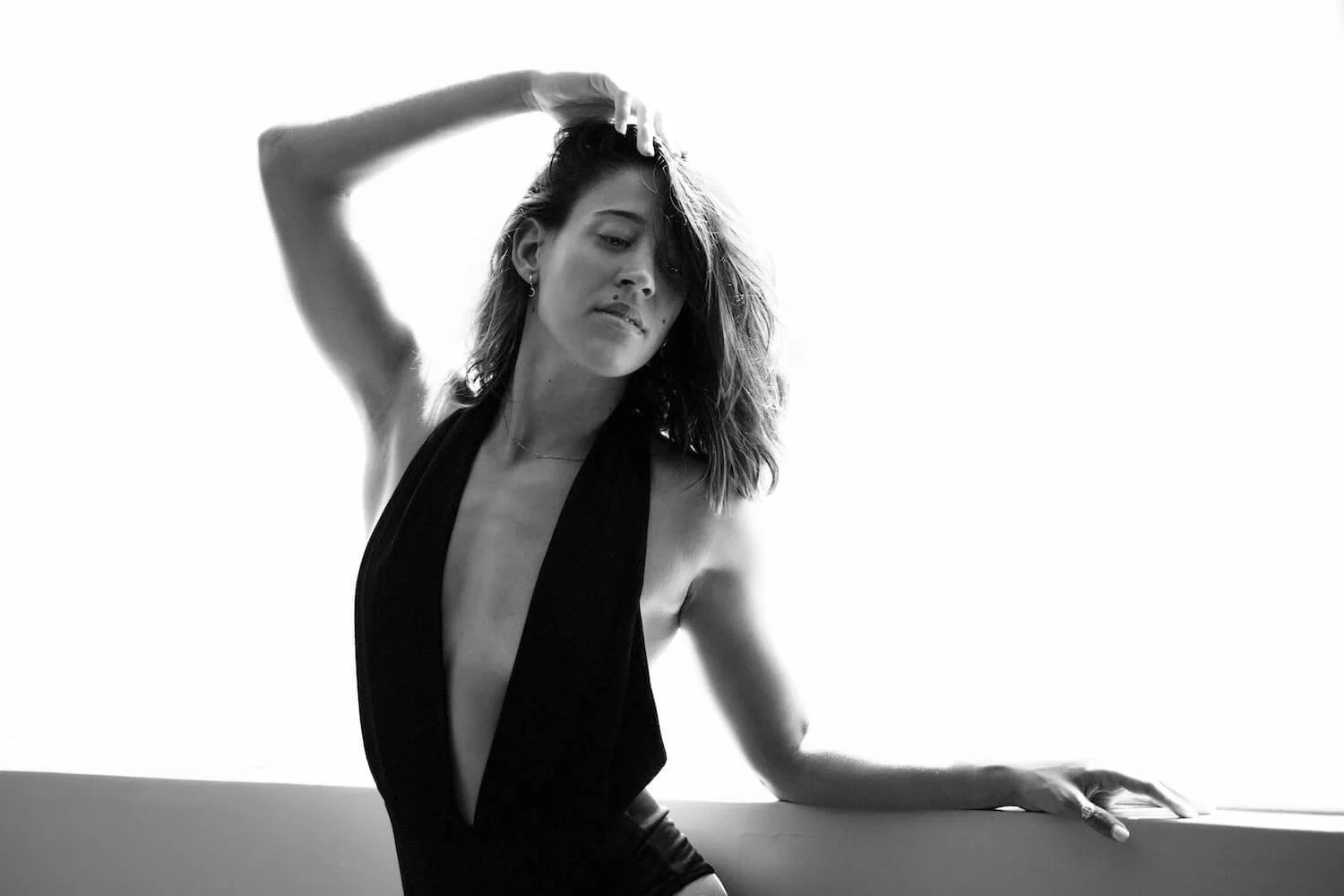 A woman with wavy hair wearing a sleeveless, deep V-neck dress, posing with her left hand on a surface and her right hand in her hair, looking down, in black and white.