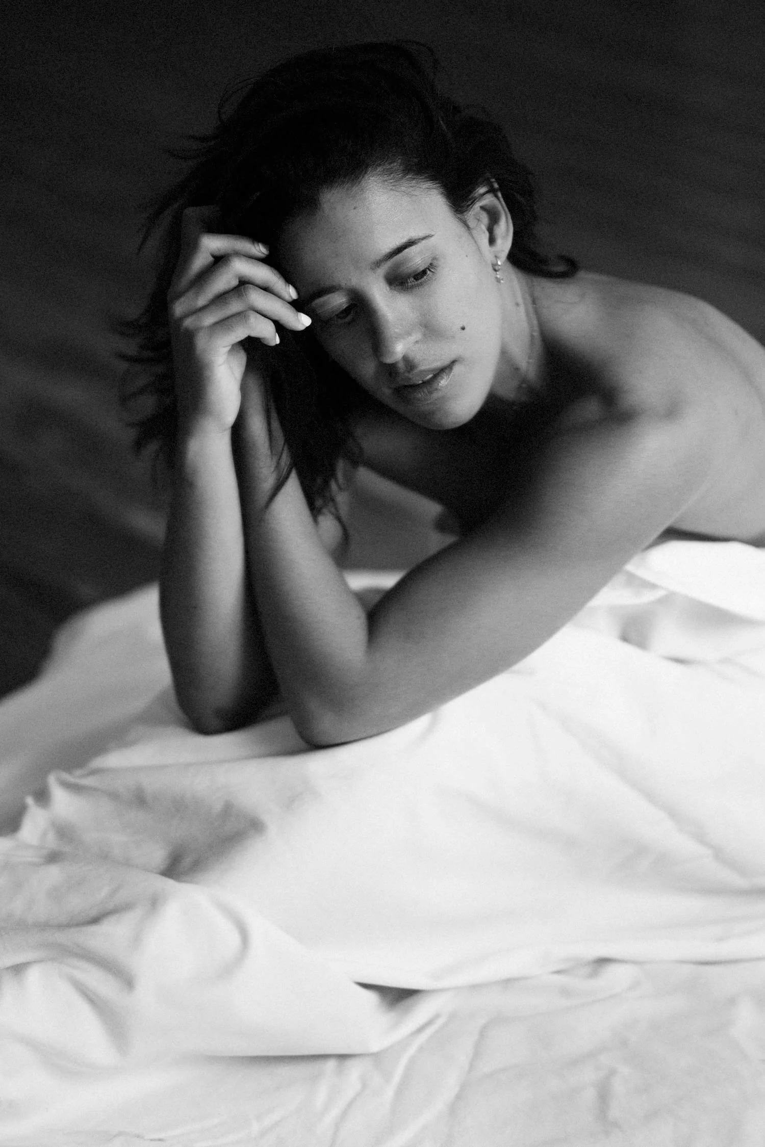 A black and white photo of a woman lying on a bed with white sheets, resting her head on her hand, with a pensive expression.