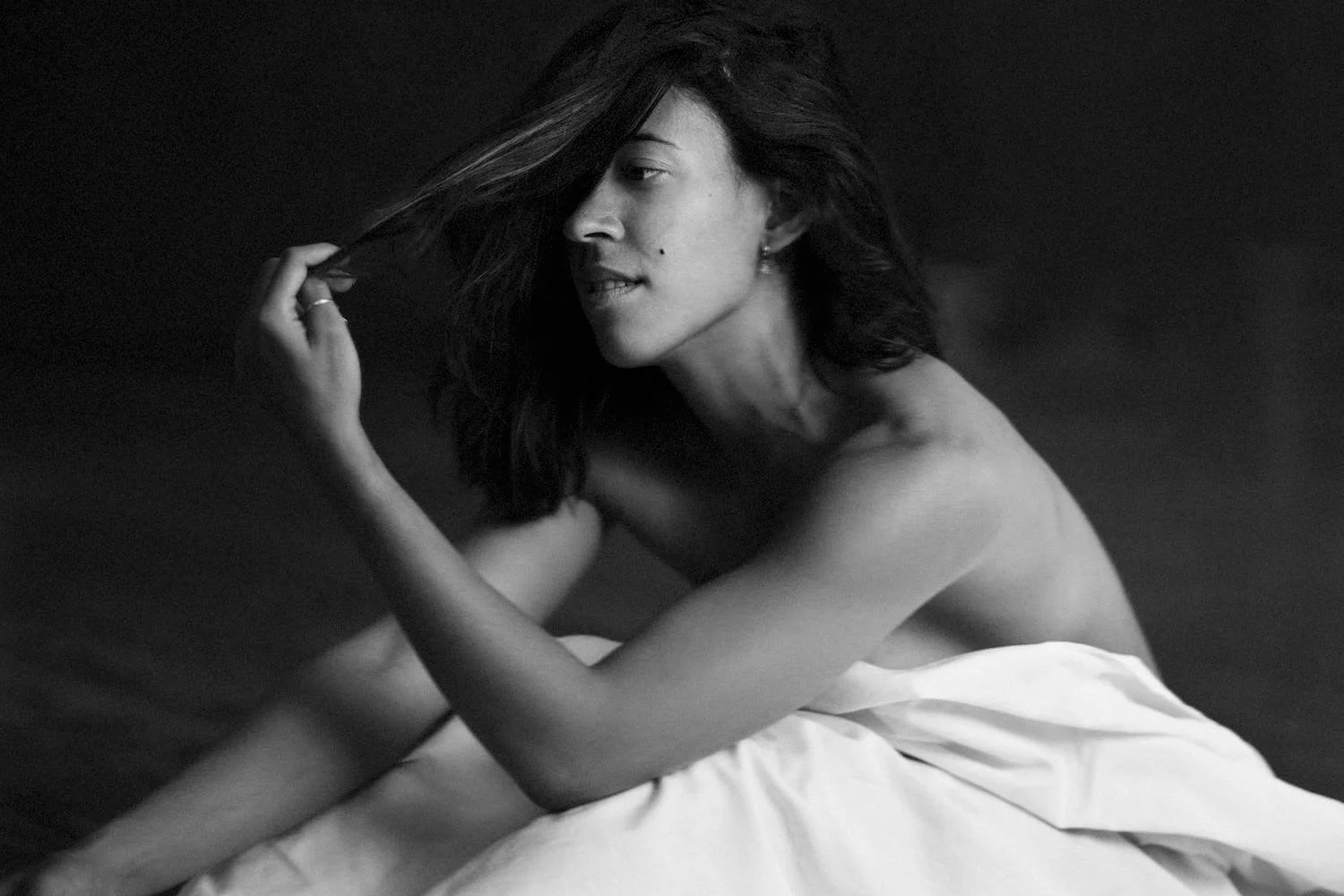 A black and white photograph of a woman with dark hair, sitting on a white sheet or blanket, topless, with her hand playing with her hair, looking to her left in a contemplative pose.