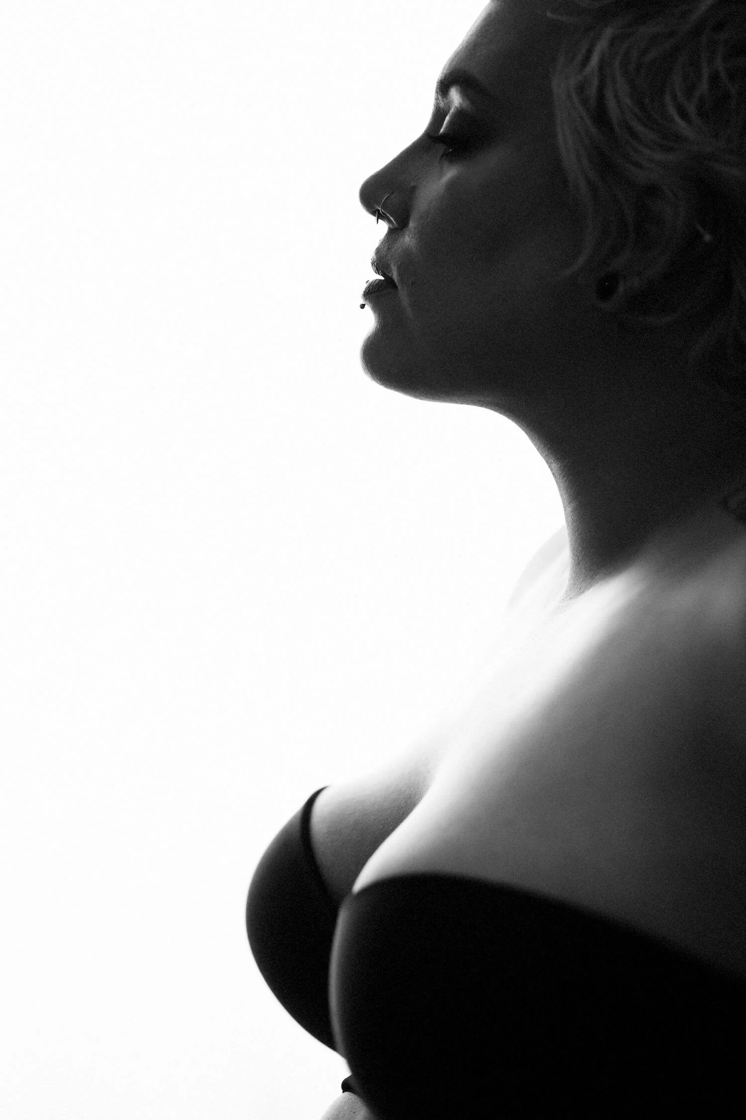 Black and white profile of a woman with short wavy hair, a septum piercing, and wearing a bra.