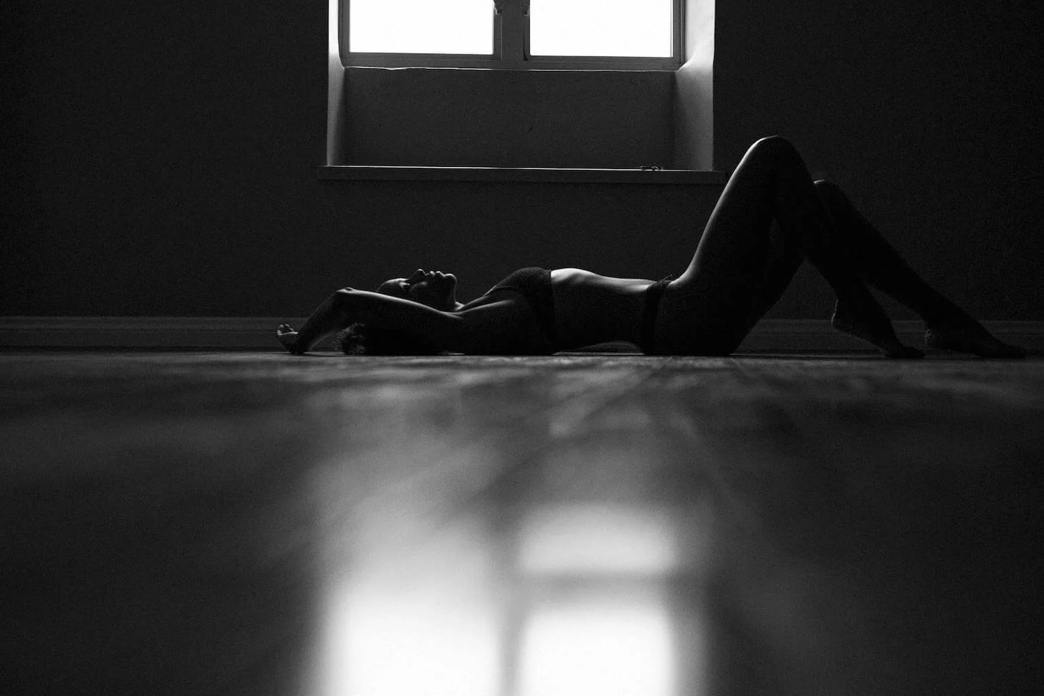 A woman lying on the floor in a dark room with a window above her, illuminated from behind, creating a silhouette effect.