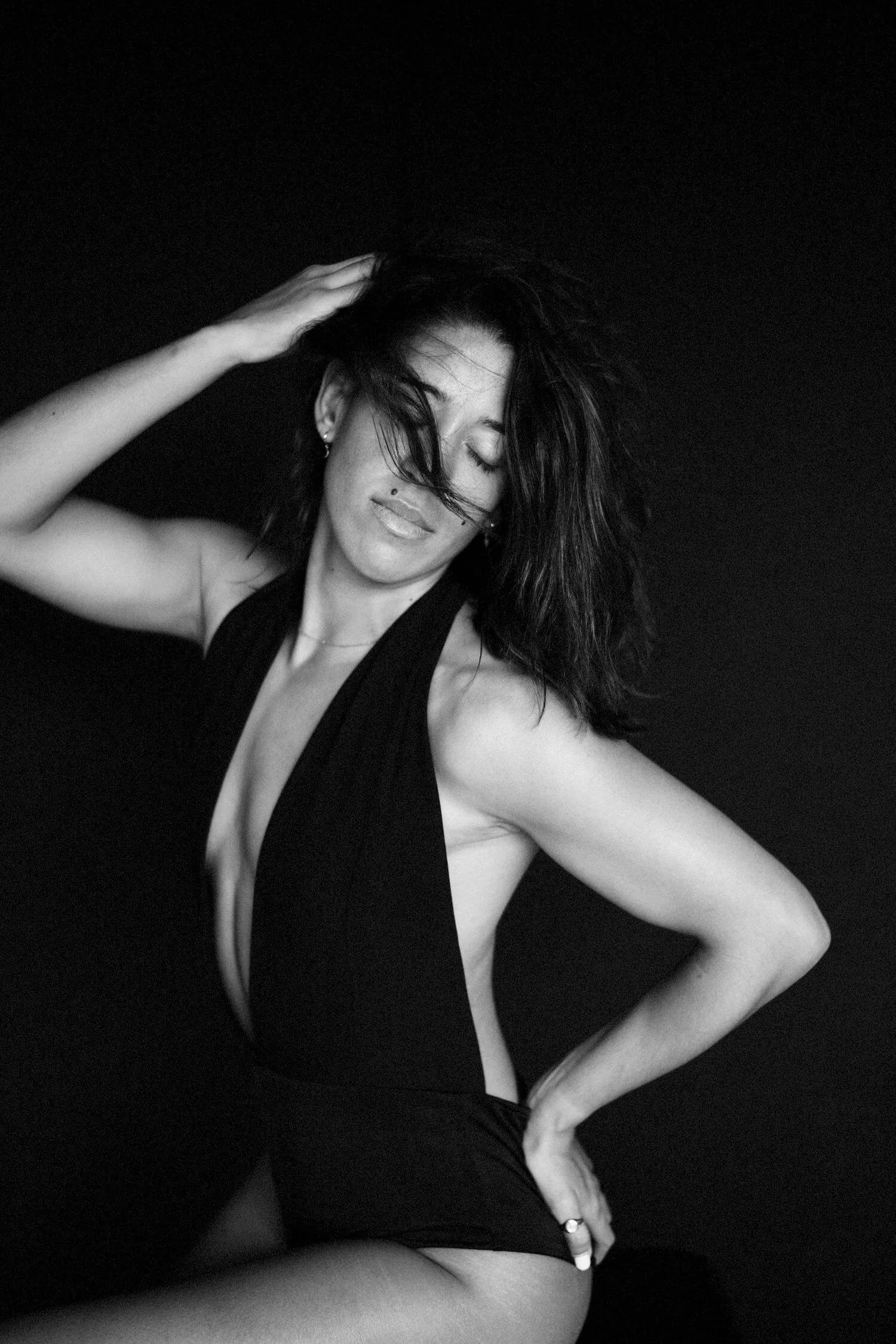 Black and white photo of a woman with shoulder-length wavy hair, wearing a sleeveless black dress with a deep neckline, posing with eyes closed against a dark background.