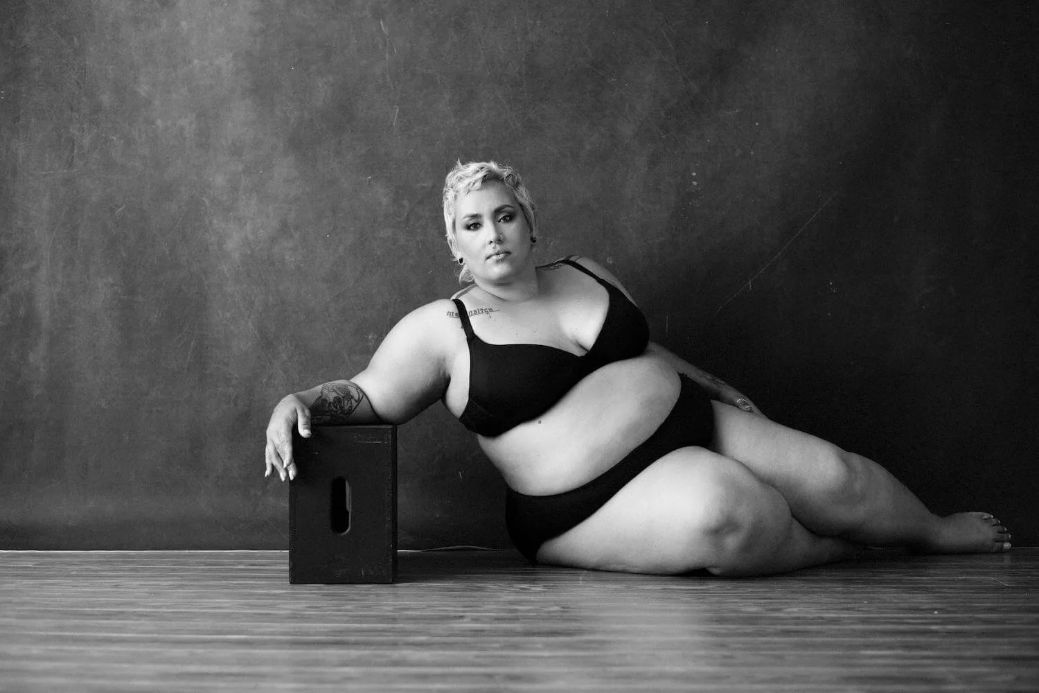 A plus-sized woman with short blonde hair, wearing black lingerie, sitting on a wooden floor with a textured wall background. She rests her left arm on a small black block, gazing at the camera with a confident expression.