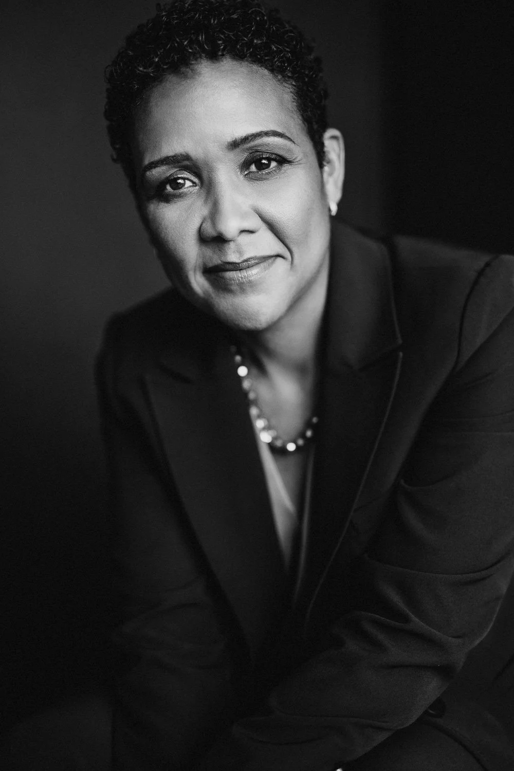 Black and white portrait of a woman with short curly hair, wearing a blazer and a pearl necklace, looking at the camera with a slight smile.