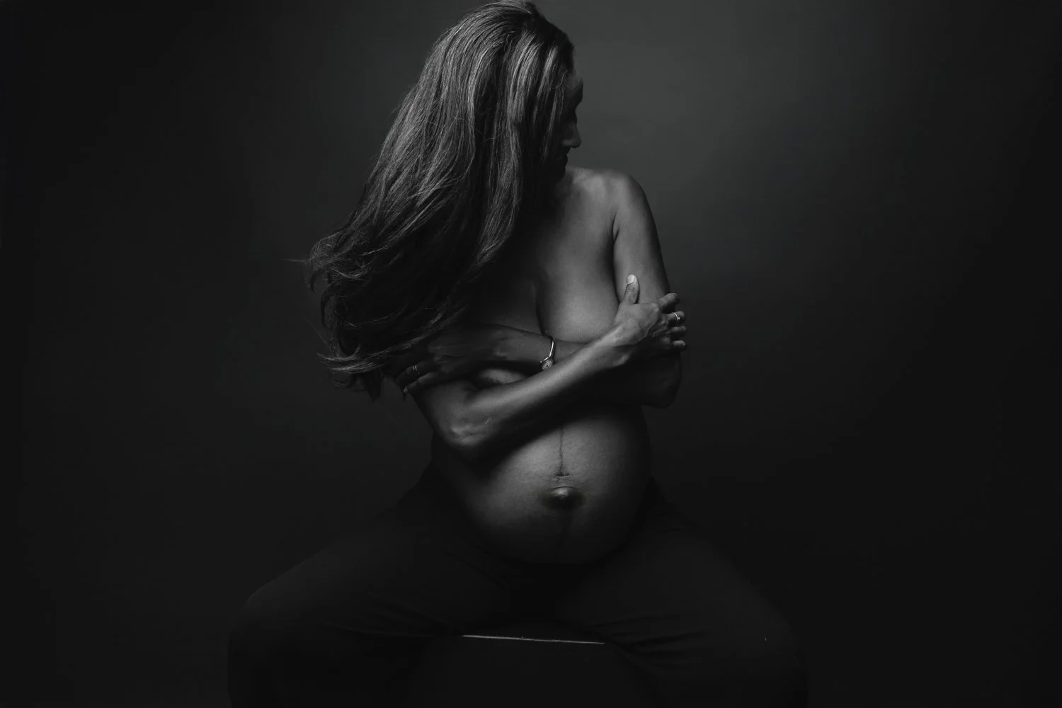 Black and white photo of a pregnant woman with long hair covering her face, topless, sitting with her arms crossed over her chest and supporting her belly.