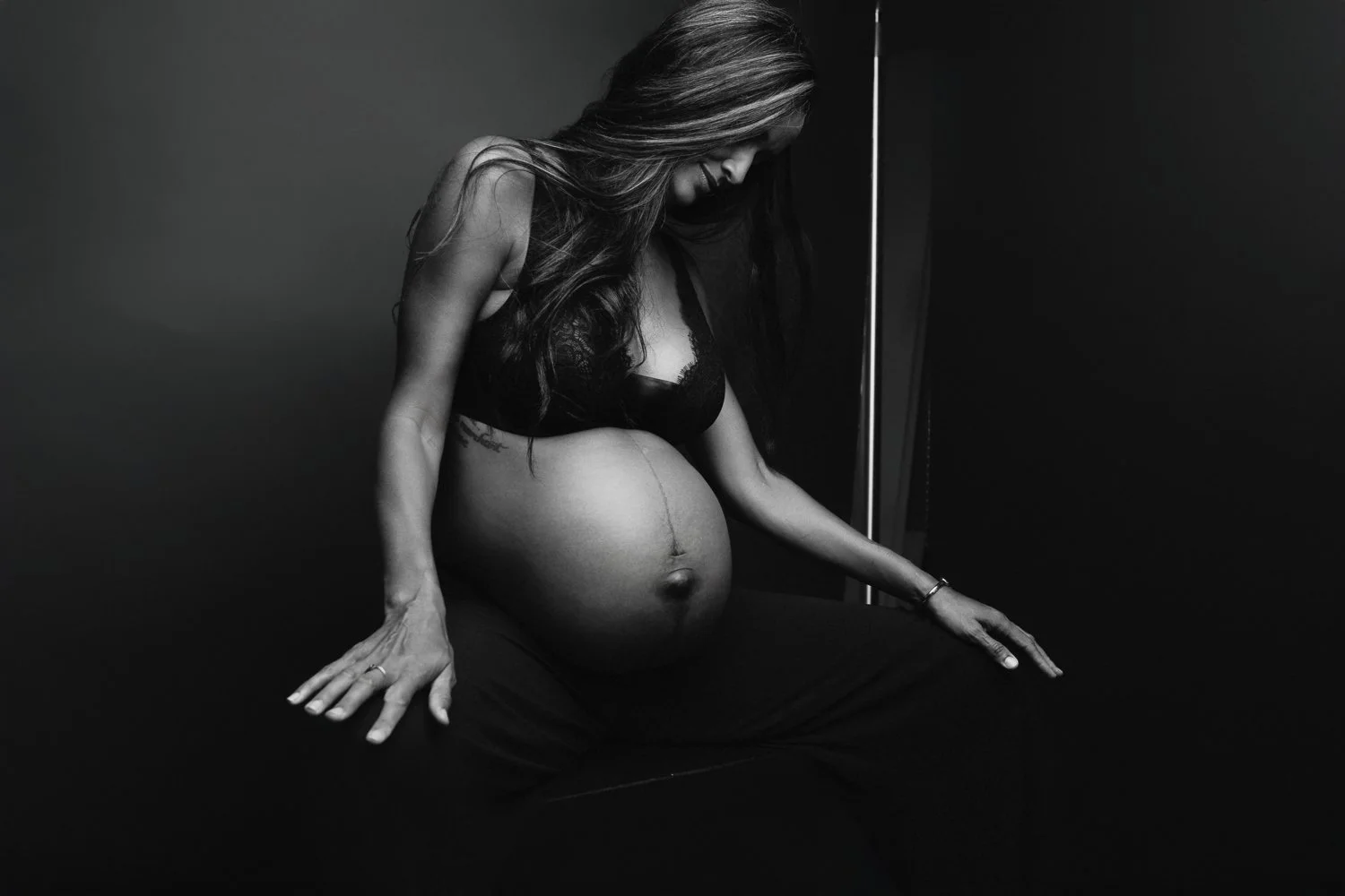 Black and white photo of a pregnant woman sitting with her head bowed, wearing a black bra and pants, with her hands resting on her lap and a visible pregnancy belly.