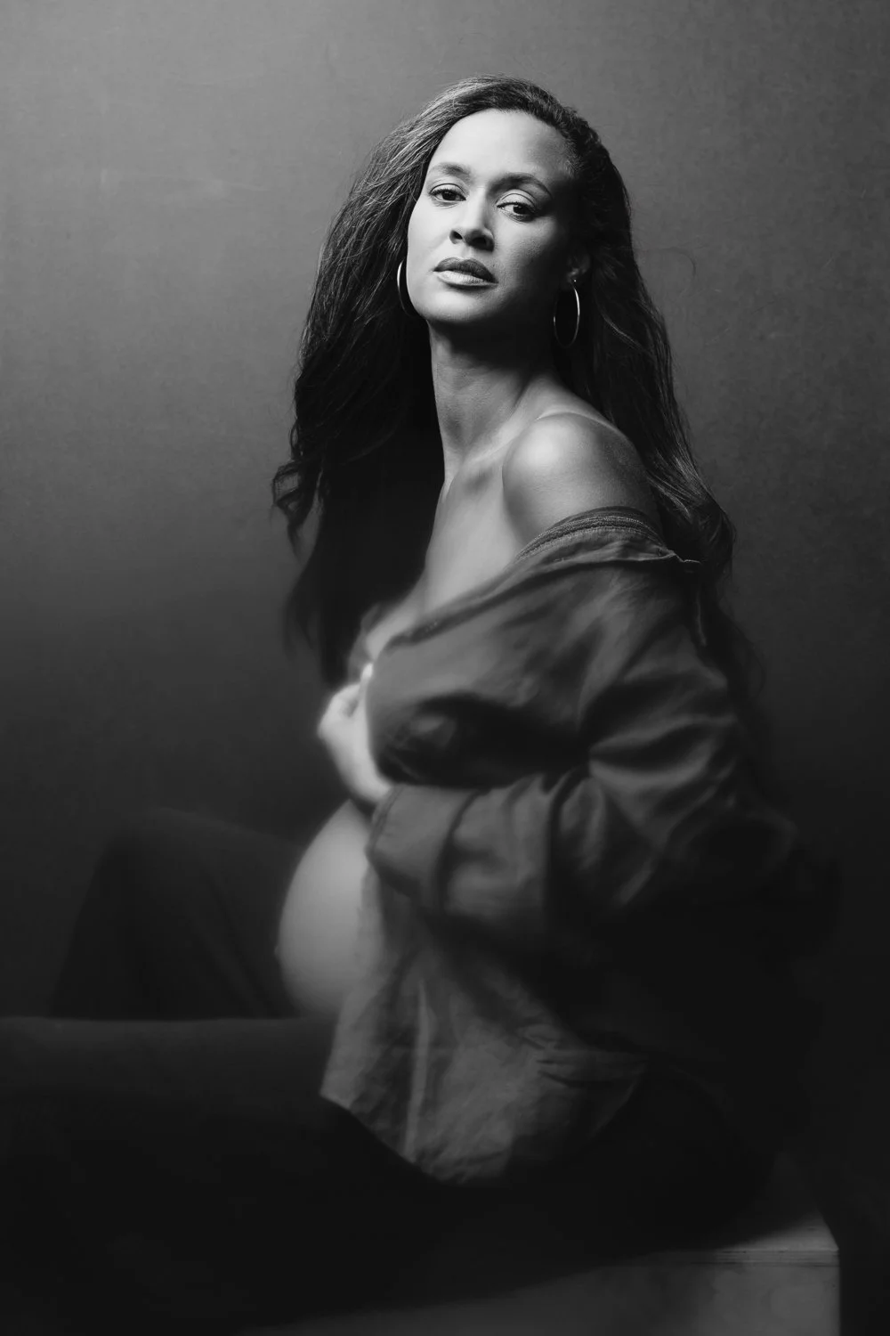 Black and white portrait of a woman with long hair, wearing hoop earrings and an open jacket, sitting against a dark background.